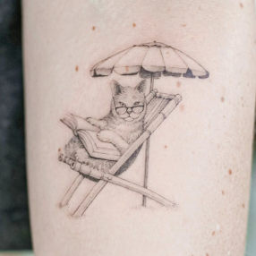 Black ink fine line tattoo of a cat wearing glasses reading a book sitting in a beach chair with an umbrella