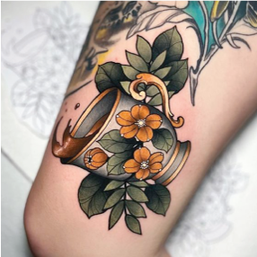A neo traditional tattoo of a grey, white, and gold tea cup surrounded by green leaves and gold flowers